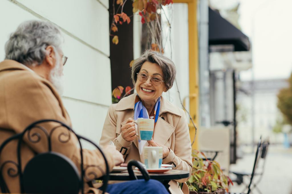 Gözlüklü ve trençkotlu neşeli son sınıf kadını, kahvaltıda kocasıyla birlikte terasta kahve içerken. - Fotoğraf, Görsel