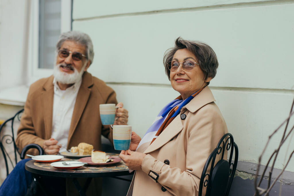 boldog idős nő szemüvegben tartó csésze, miközben villásreggeli szakállas férj a teraszon  - Fotó, kép