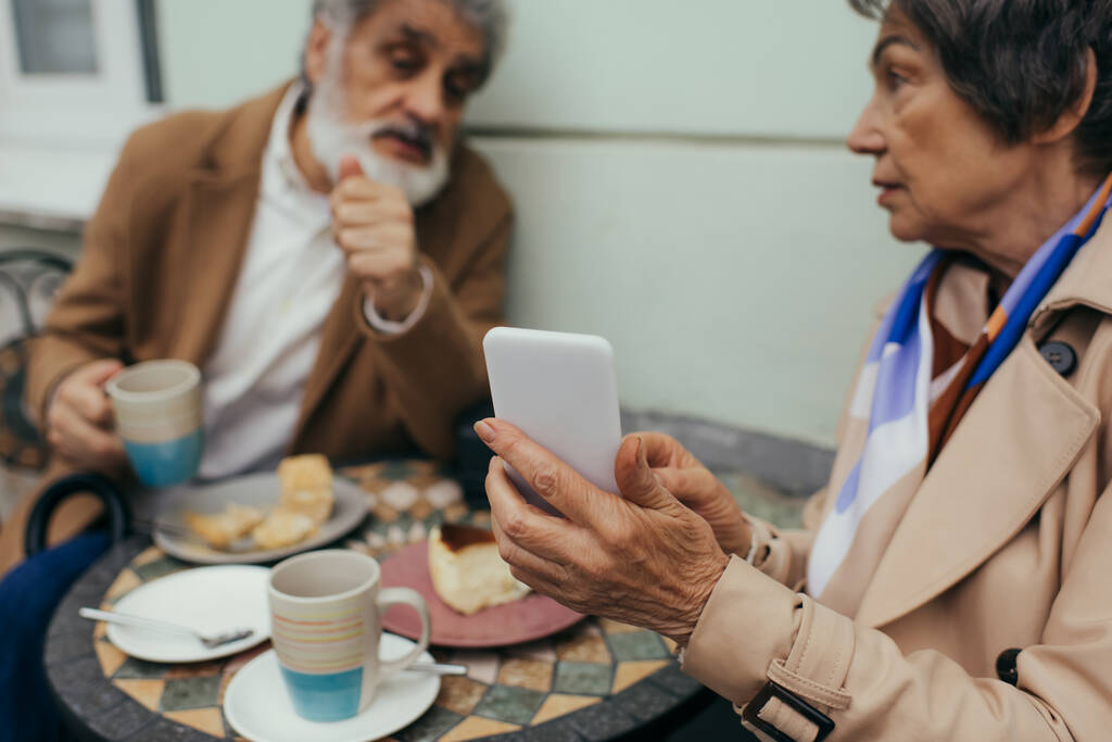 vanhempi nainen tilalla älypuhelin kun ottaa brunssi miehensä terassilla kahvila - Valokuva, kuva