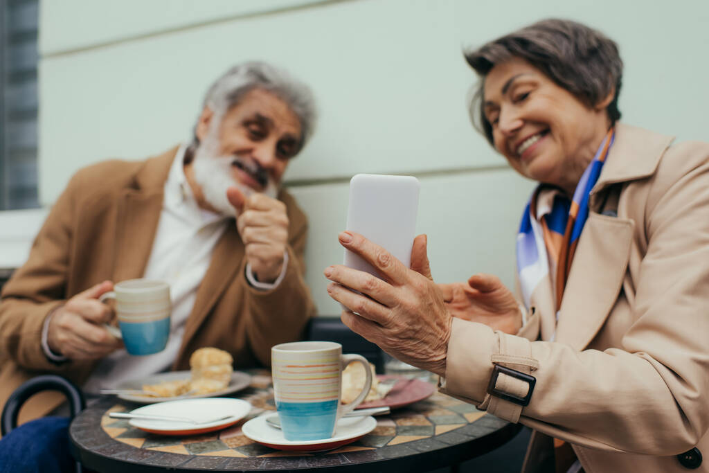 щаслива старша жінка тримає смартфон, маючи бранч з чоловіком на терасі кафе
 - Фото, зображення
