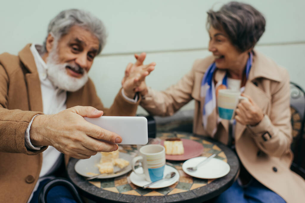 verschwommene und glückliche Senioren beim Videoanruf während des Frühstücks auf der Terrasse des Cafés  - Foto, Bild