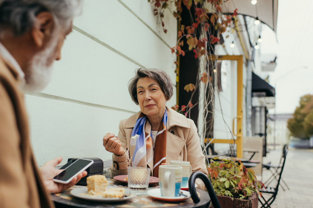 ευτυχισμένη ηλικιωμένη γυναίκα τρώει κέικ και ψάχνει στο σύζυγο με smartphone στη βεράντα του καφέ - Φωτογραφία, εικόνα