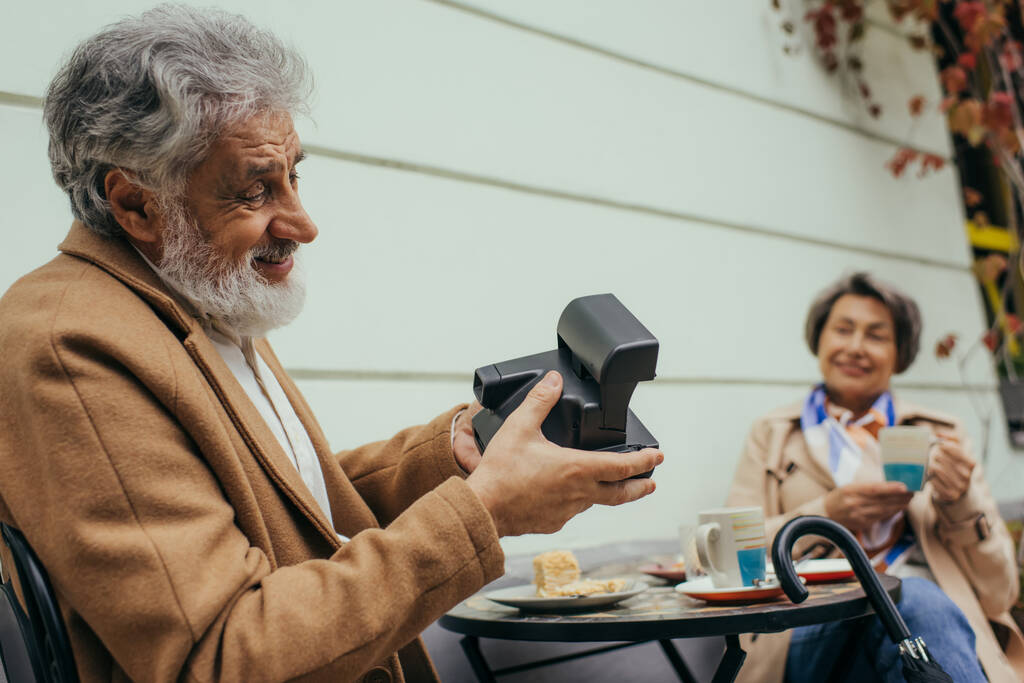 felice uomo barbuto in possesso di fotocamera vintage e scattare foto di moglie anziana offuscata durante la colazione  - Foto, immagini