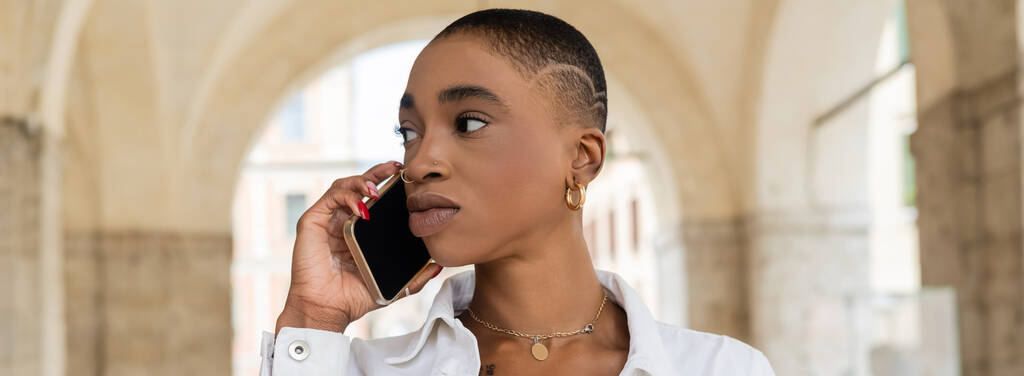 Kısa saçlı Afrikalı Amerikalı kadın Treviso 'daki bulanık şehir sokağında akıllı telefondan konuşuyor.  - Fotoğraf, Görsel