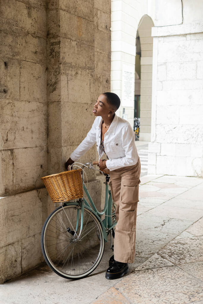 Femme afro-américaine aux cheveux courts debout près du vélo dans la rue urbaine en Italie - Photo, image