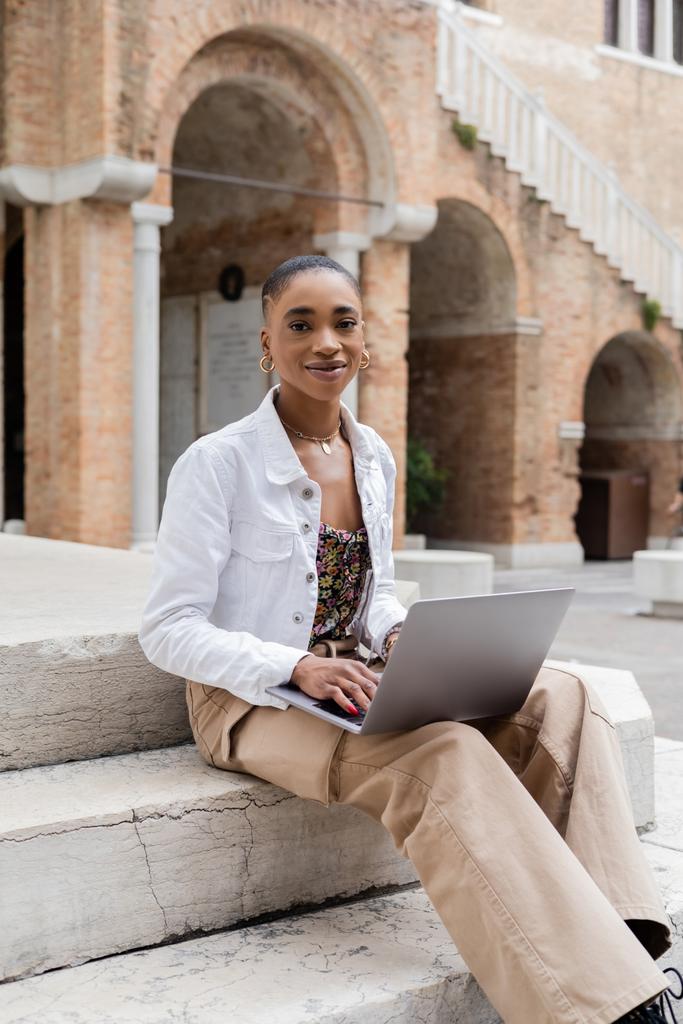 Positive african american blogger using laptop and looking at camera on stairs outdoors in Italy - Photo, Image