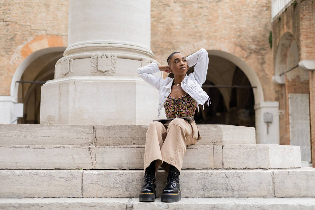 Freelance afro-américaine aux cheveux courts avec ordinateur portable assis sur les escaliers dans la rue de la ville en Italie - Photo, image