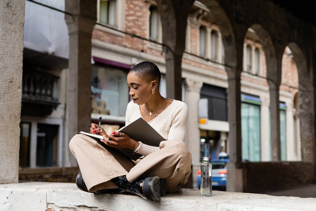 Mujer afroamericana de moda escribiendo en un cuaderno cerca de una botella de agua en la calle urbana de Treviso - Foto, Imagen