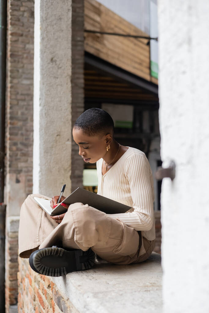 Fashionable Afrikaans-Amerikaanse vrouw schrijven op notebook in de boog van het gebouw buiten in Treviso - Foto, afbeelding