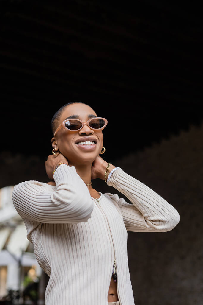 Tiefansicht einer trendigen afrikanisch-amerikanischen Frau mit Sonnenbrille, die auf der Stadtstraße lächelt  - Foto, Bild