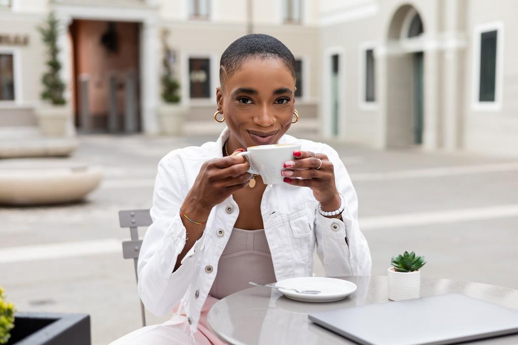 Стильный африканский фрилансер держит чашку кофе возле ноутбука и растение в открытом кафе в Италии - Фото, изображение