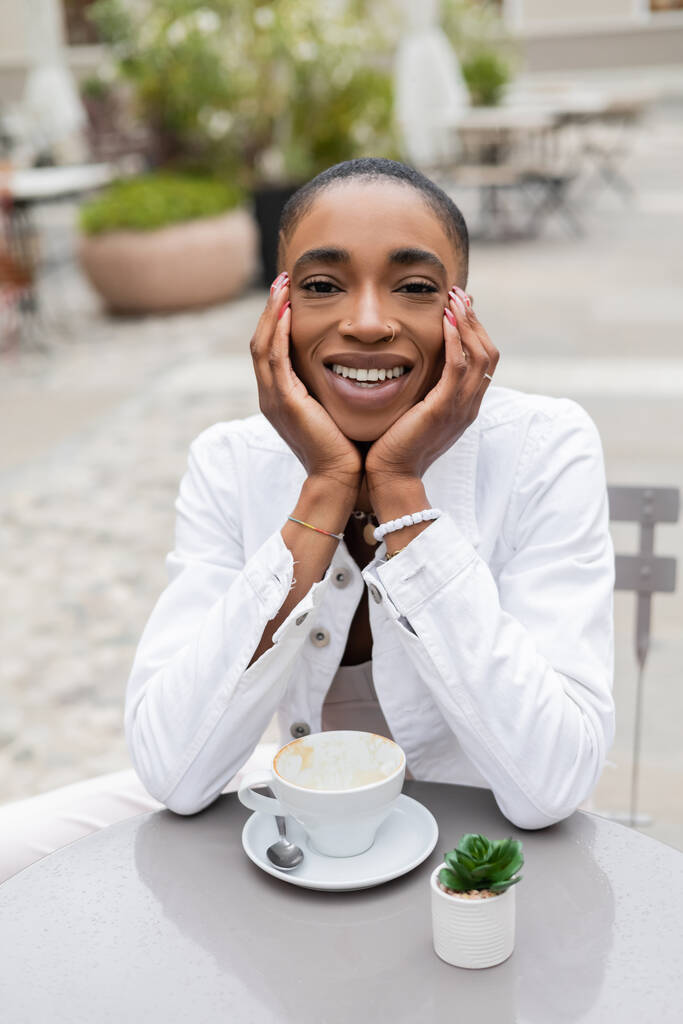 Happy african american woman looking at camera near cup and plant in outdoor cafe  - Photo, Image