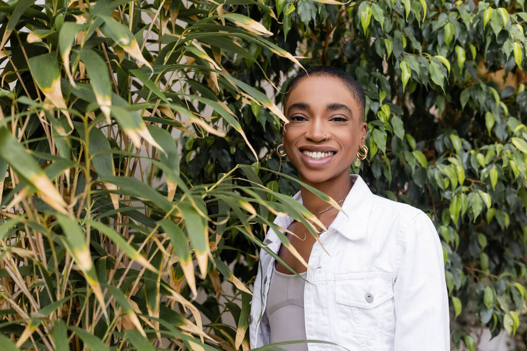Sonriente mujer afroamericana de pelo corto mirando a la cámara cerca de las plantas al aire libre  - Foto, Imagen