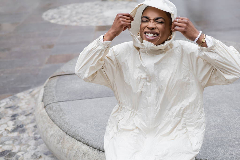Ευτυχισμένη και κομψή Αφροαμερικανή με αδιάβροχο που κλείνει τα μάτια στον αστικό δρόμο.  - Φωτογραφία, εικόνα