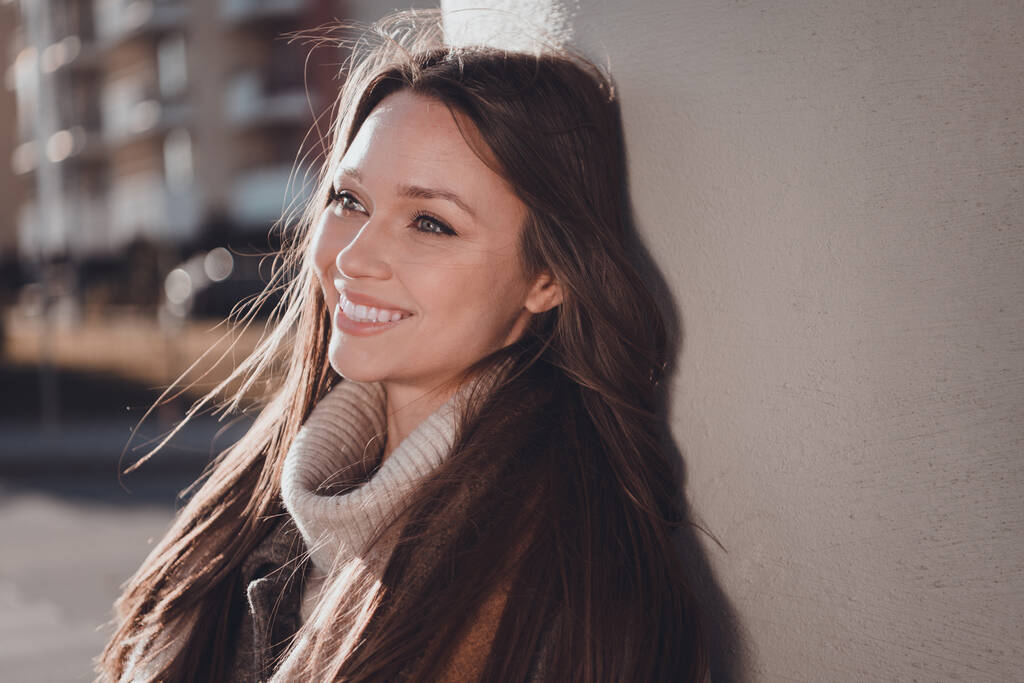 Zdjęcie uroczej uroczej kobiety w szarym płaszczu uśmiechniętej cieszącej się porannym słońcem poza ulicą miejską. - Zdjęcie, obraz