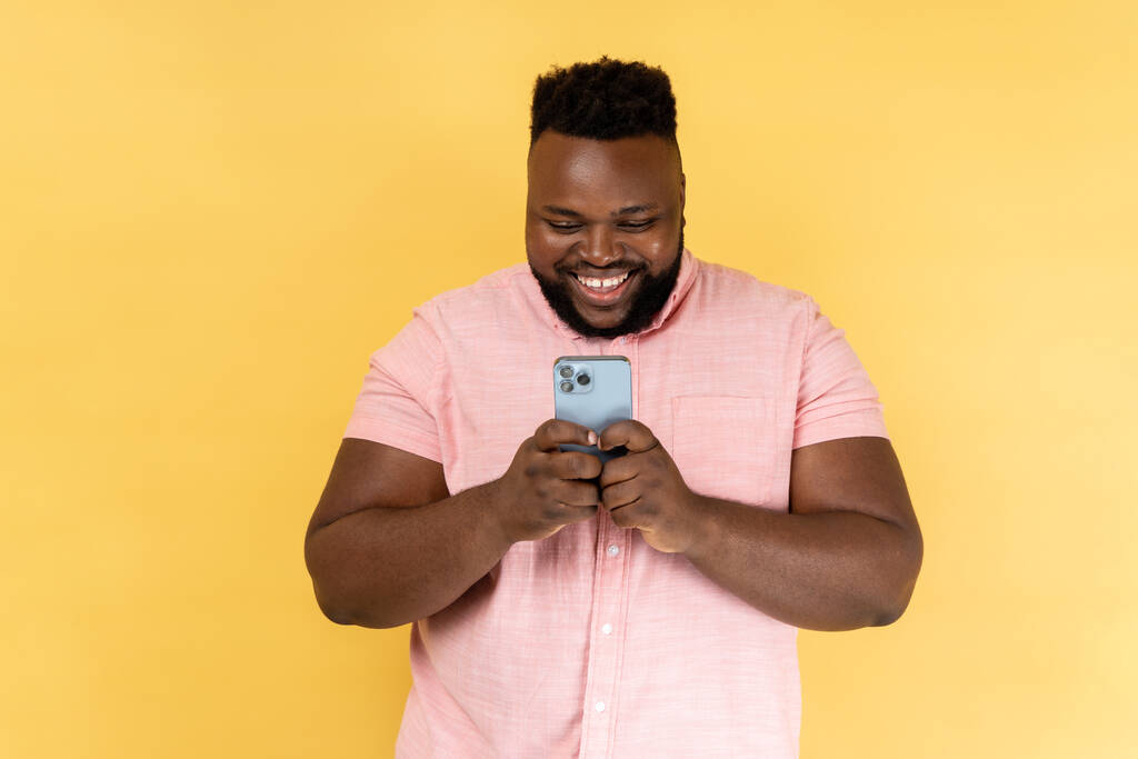 Portrét vousatého šťastného muže v růžové košili, který používá mobilní telefon s veselým výrazem, závislý na smartphonu, textuje na sociální síti. Vnitřní studio záběr izolované na žlutém pozadí. - Fotografie, Obrázek
