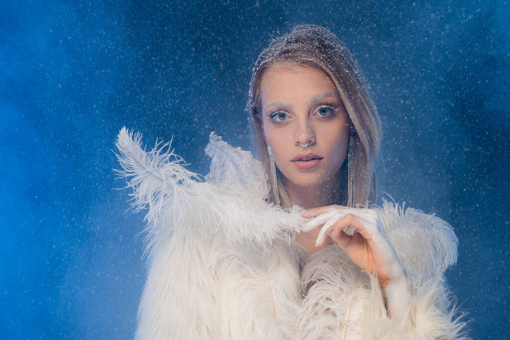 mujer joven con maquillaje de invierno sosteniendo pluma blanca bajo la nieve que cae en azul oscuro  - Foto, Imagen