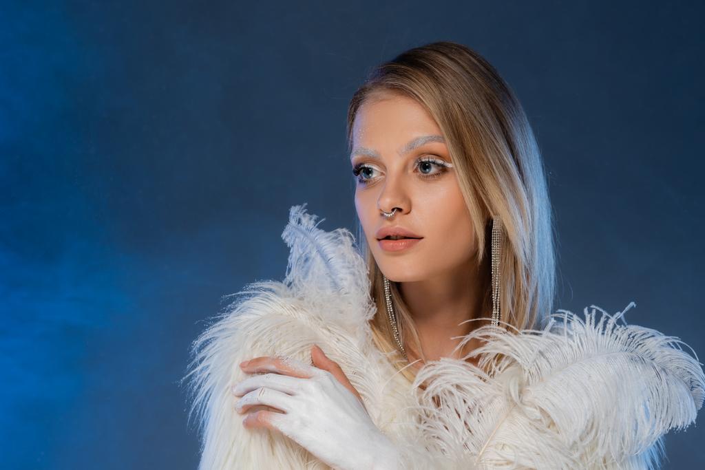 blond vrouw met winter make-up houden witte veer op donkerblauwe achtergrond - Foto, afbeelding