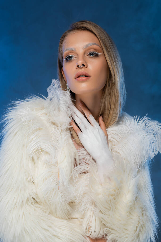 mujer joven perforada con maquillaje de invierno posando en chaqueta de piel sintética blanca con plumas en azul oscuro  - Foto, imagen