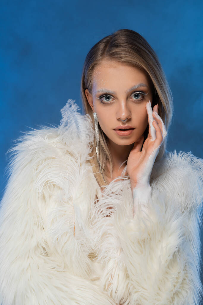 przekłuta młoda kobieta z makijażem zimowym pozowanie w białym futra faux kurtka i patrząc na aparat fotograficzny na granatowym  - Zdjęcie, obraz