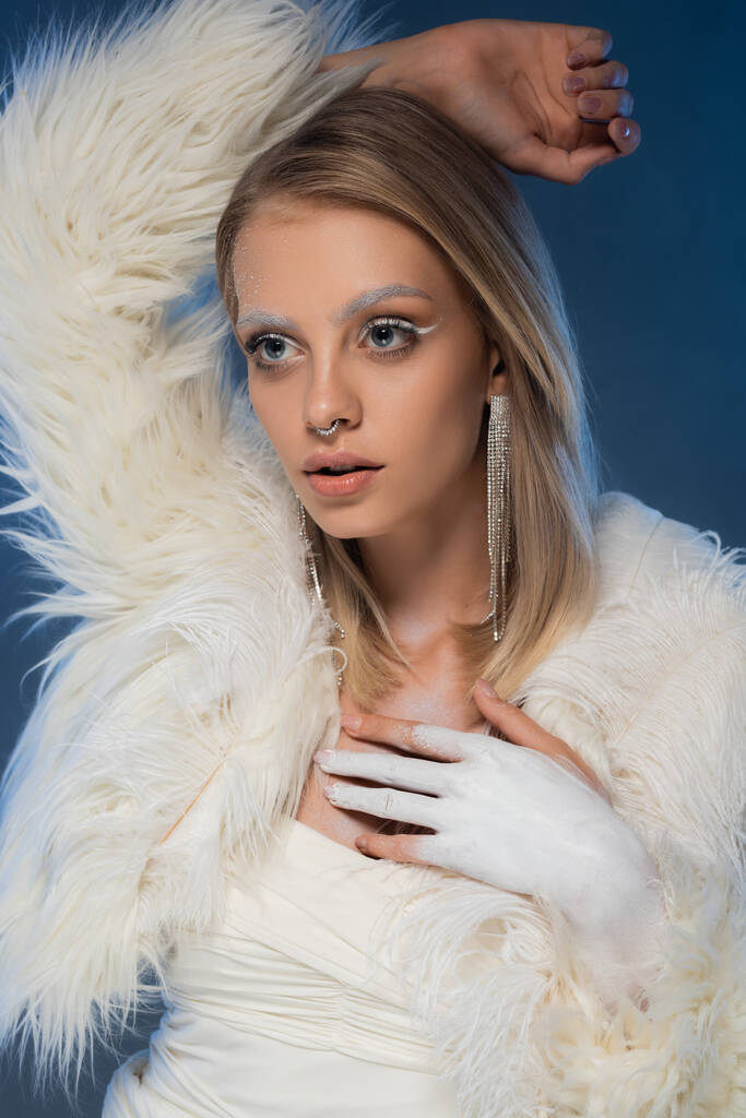 pierced young woman with winter makeup posing in white jacket with feathers on dark blue  - Photo, Image