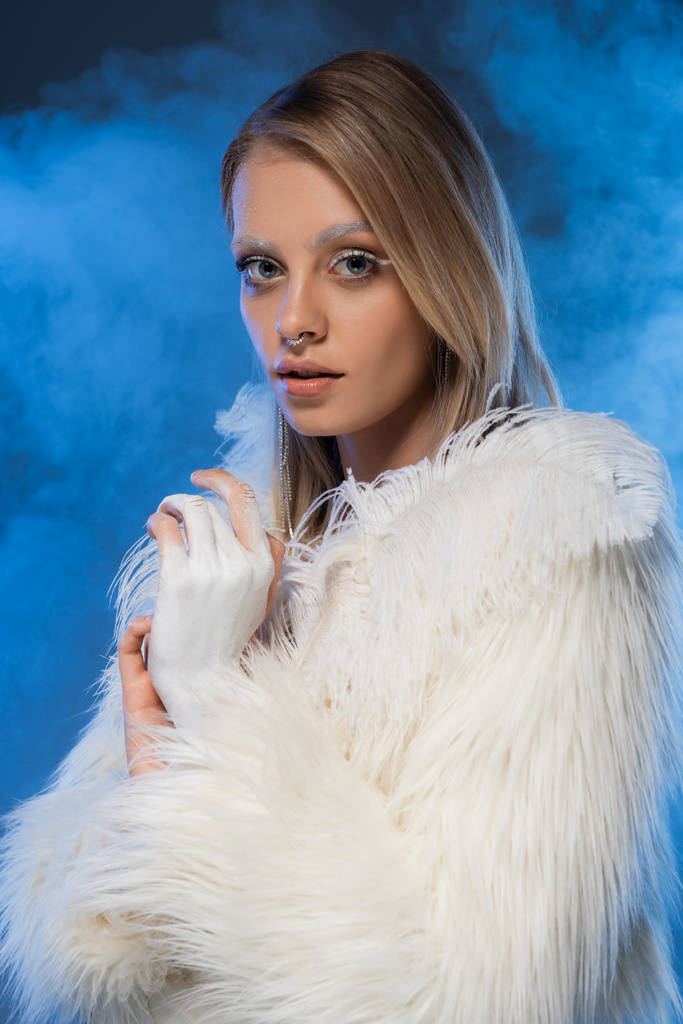 donna traforata con trucco invernale in posa in giacca di pelliccia sintetica con piume su blu con fumo - Foto, immagini