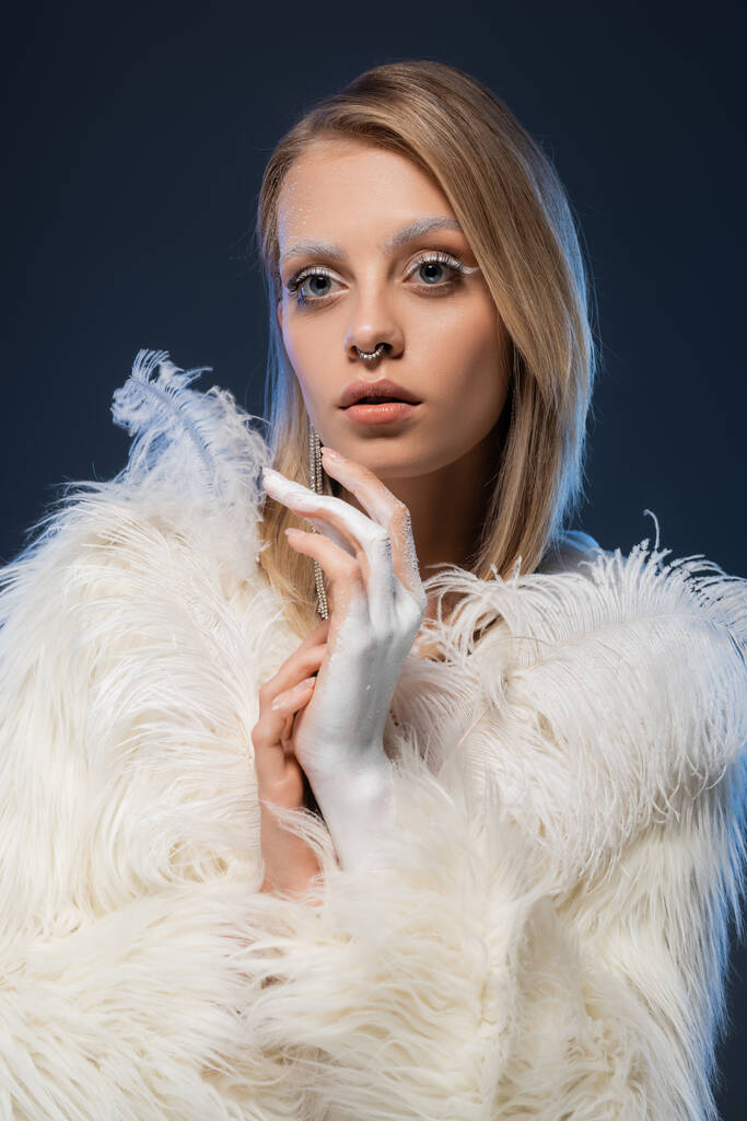 modelo joven perforado con maquillaje de invierno posando en chaqueta de piel sintética blanca con plumas aisladas en azul oscuro  - Foto, imagen