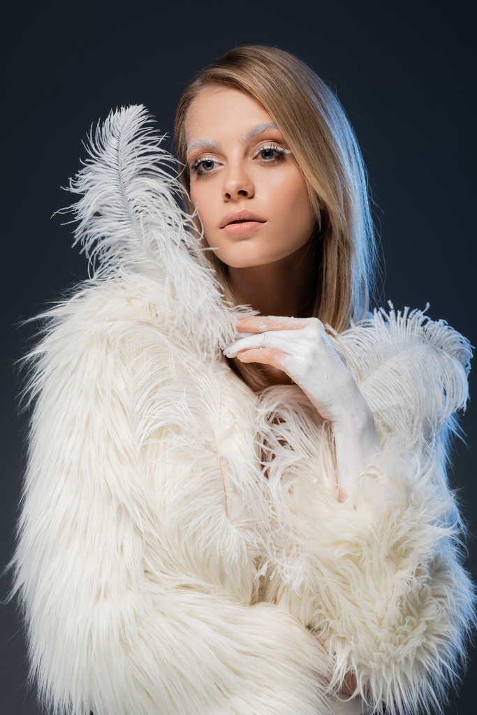 pretty young woman in white faux fur jacket posing with feather isolated on blue - Photo, Image