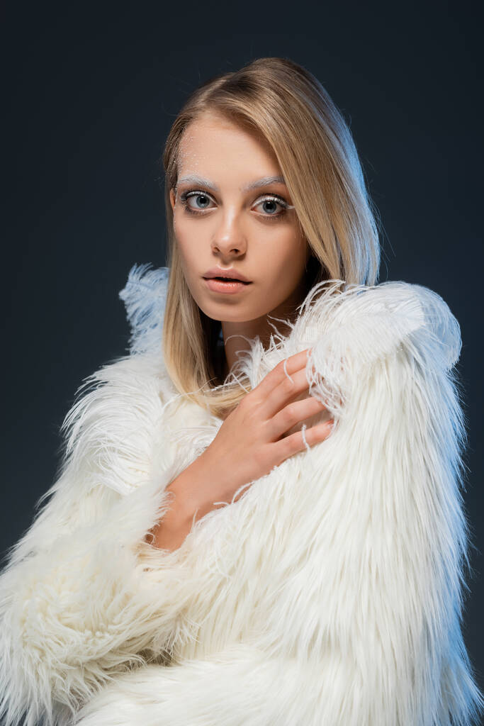 young blonde woman with winter makeup looking at camera and posing in faux fur jacket isolated on blue - Photo, Image
