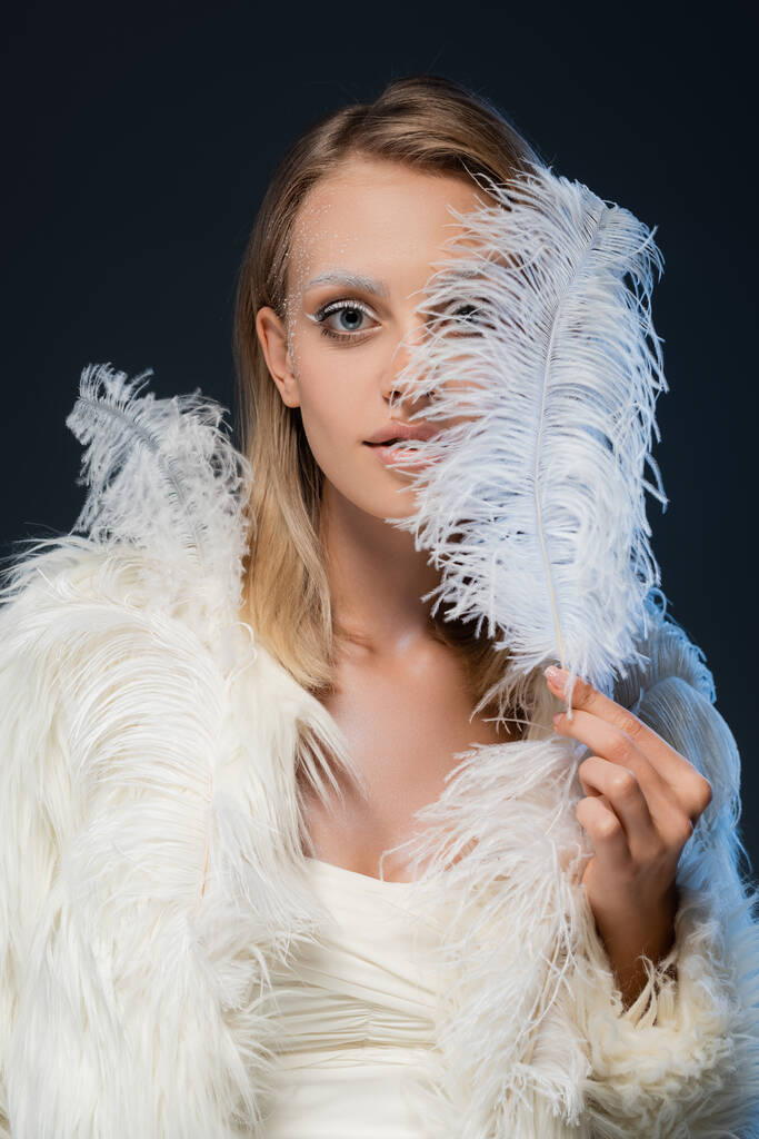 jeune femme blonde avec maquillage d'hiver regardant la caméra couvrant le visage avec plume isolé sur bleu - Photo, image