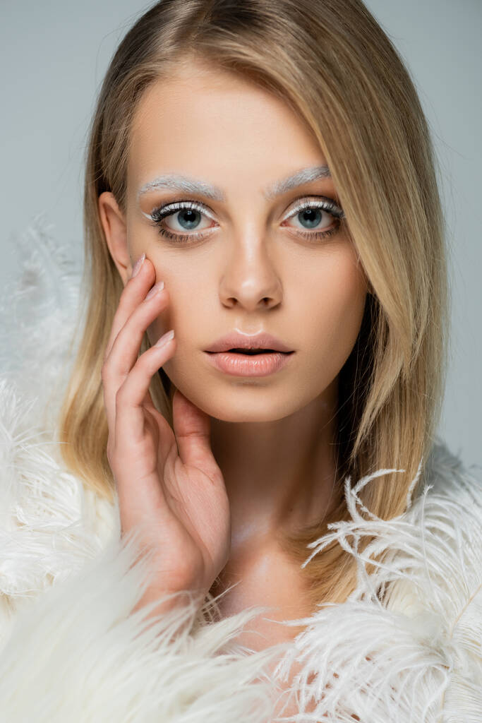 young model in winter faux fur jacket with feathers looking at camera while touching face isolated on grey - Photo, Image