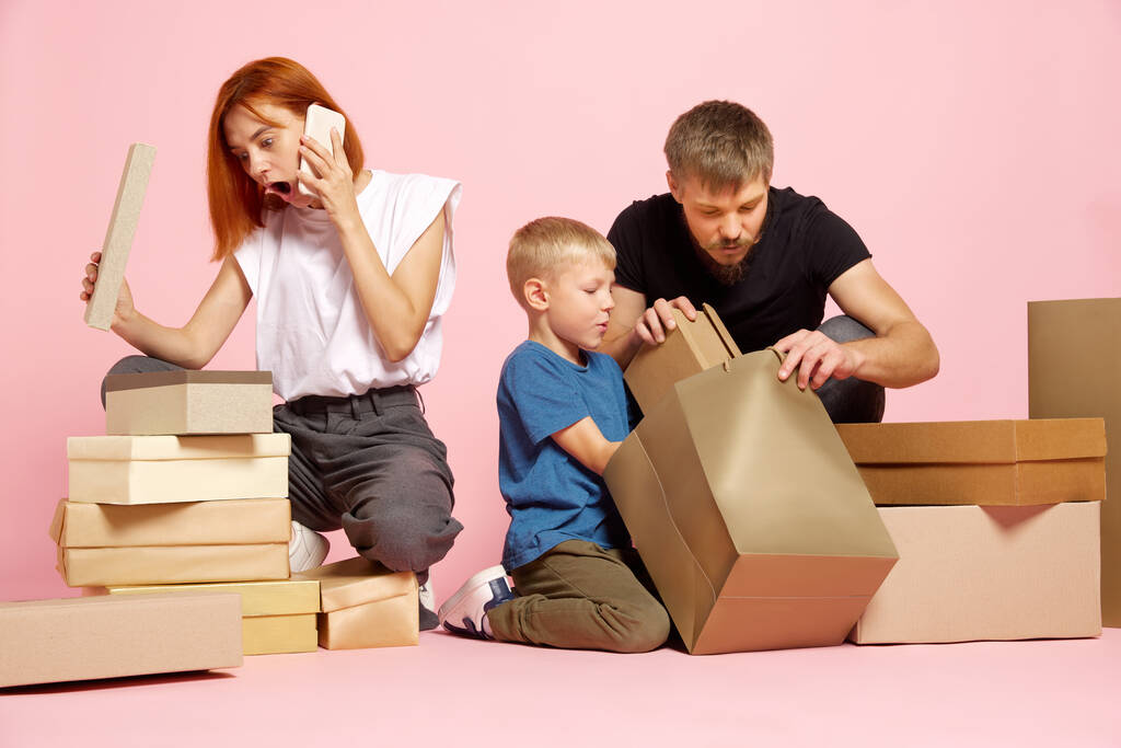 En tiedä. Onnellinen äiti, isä ja poika avaavat pahvilaatikoita, saavat paketteja purkamisen jälkeen verkkokaupoissa vaaleanpunaisen studion taustalla. Pura paketti, postitoimitus ja iso myyntipäivä käsite - Valokuva, kuva