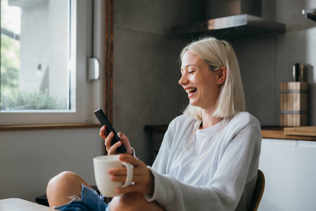 lachende gen z vrouw drinken koffie en het gebruik van mobiele telefoon in haar huis - Foto, afbeelding