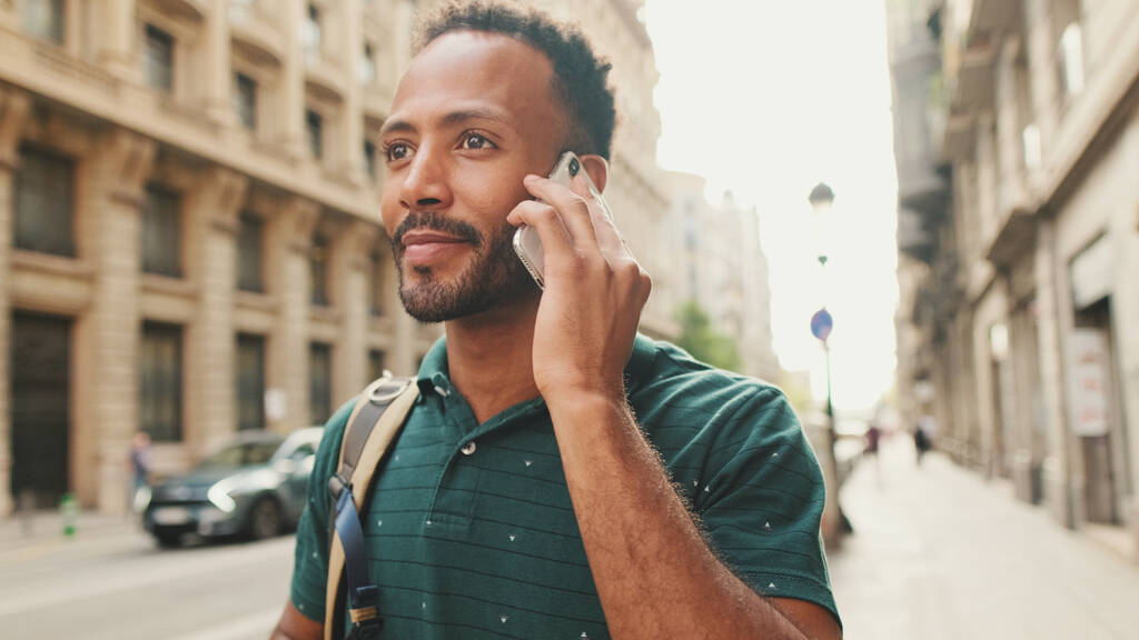 Κοντινό πλάνο, νεαρός άνδρας περπατά στο δρόμο στην παλιά πόλη και περνώντας αυτοκίνητα στο παρασκήνιο και μιλάει στο τηλέφωνο - Φωτογραφία, εικόνα