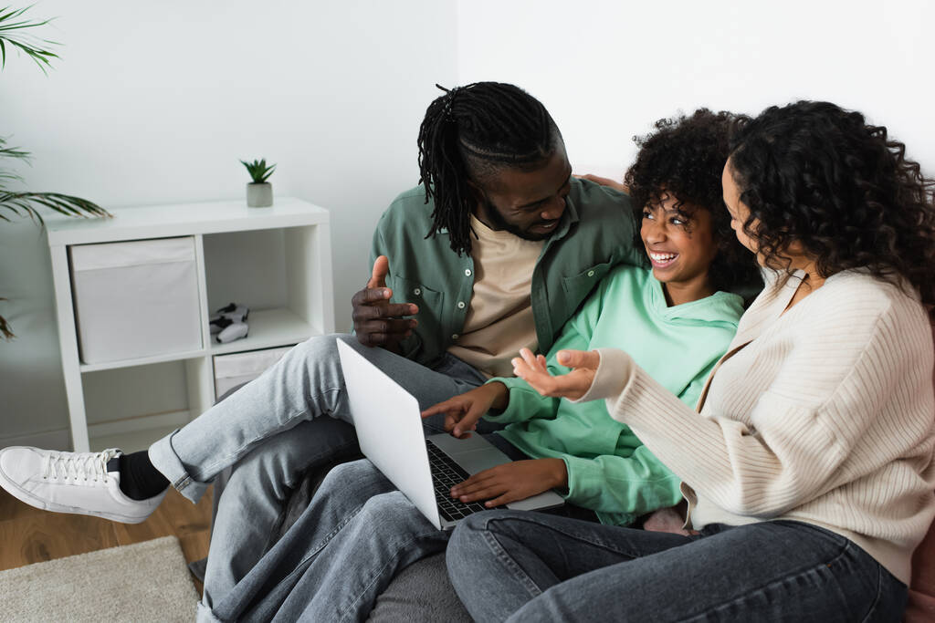 щасливі афроамериканські батьки дивляться на усміхнену дев'ятнадцять доньку з ноутбуком, що сидить на дивані
 - Фото, зображення