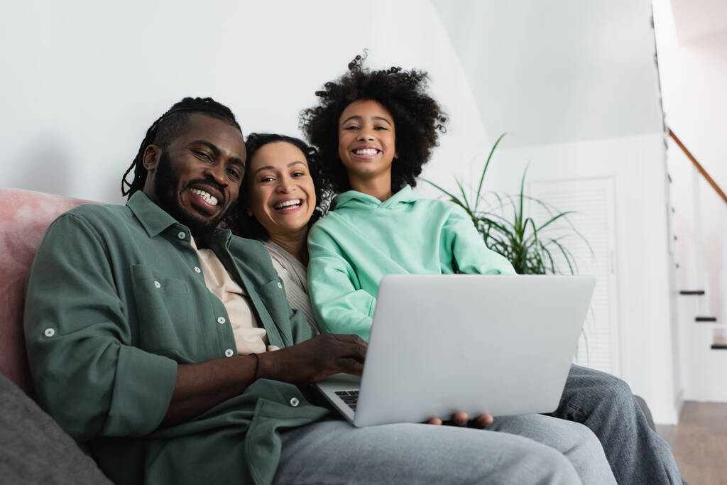 χαρούμενη αφρικάνικη αμερικανική οικογένεια κοιτάζοντας κάμερα κοντά στο φορητό υπολογιστή στο σαλόνι  - Φωτογραφία, εικόνα