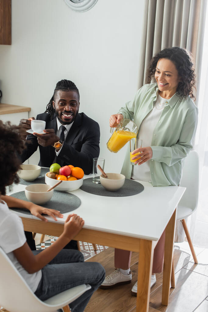 陽気なアフリカ系アメリカ人女性スーツ姿の幸せな夫と娘の近くのガラスにオレンジジュースを注ぐ朝食中に  - 写真・画像