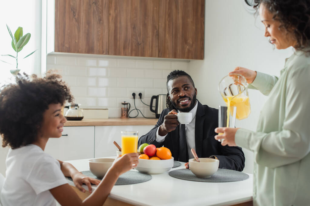 Χαρούμενος Αφροαμερικάνος επιχειρηματίας με κοστούμι κοιτάζοντας τη σύζυγο να ρίχνει χυμό πορτοκάλι σε ποτήρι κατά τη διάρκεια του πρωινού  - Φωτογραφία, εικόνα