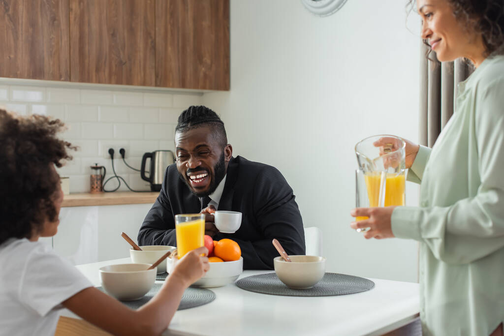 Χαρούμενος Αφροαμερικάνος επιχειρηματίας με κοστούμι να γελάει και να κοιτάζει την κόρη του κοντά στη σύζυγο με μια κανάτα χυμό πορτοκάλι. - Φωτογραφία, εικόνα