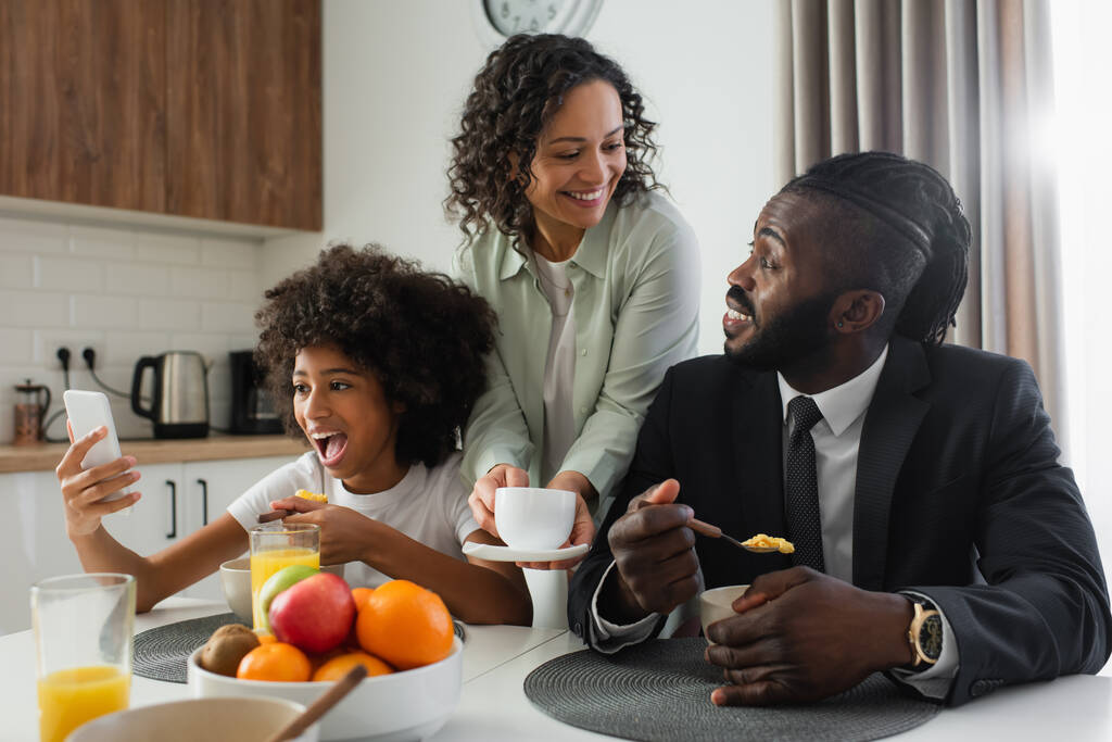 mulher americana africana alegre segurando xícara de café perto do marido de terno enquanto a filha pré-adolescente usando smartphone  - Foto, Imagem