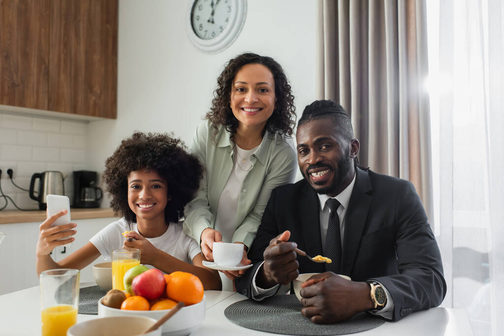 allegra donna afroamericana che tiene in mano una tazza di caffè vicino al marito in giacca e cravatta e figlia preadolescente con smartphone  - Foto, immagini