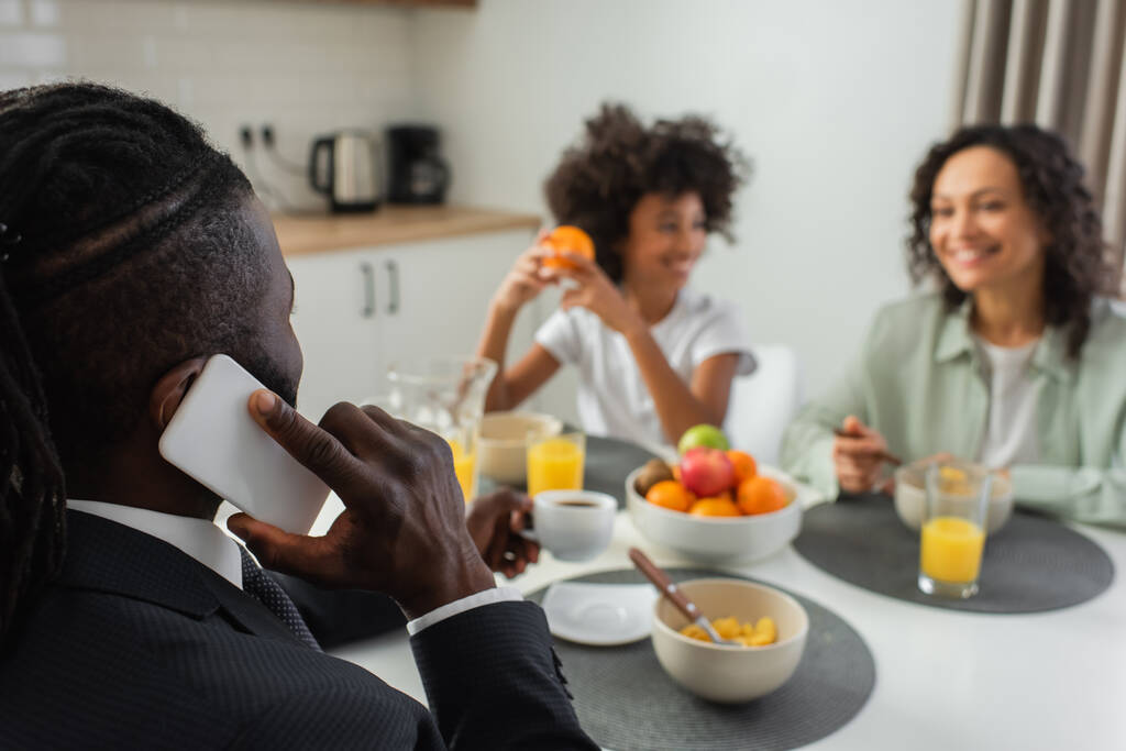africký americký podnikatel mluví na smartphone během snídaně s rodinou na rozmazaném pozadí - Fotografie, Obrázek