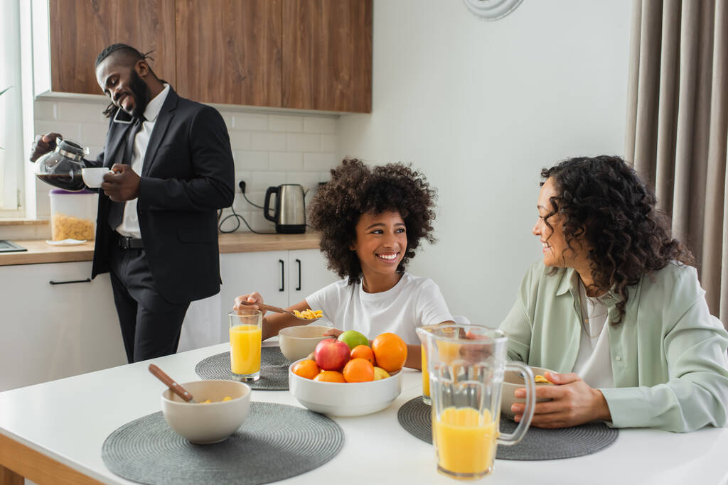szczęśliwy Afryki amerykańska matka i córka patrząc na siebie w pobliżu biznesmen mówi na smartfona podczas śniadania  - Zdjęcie, obraz