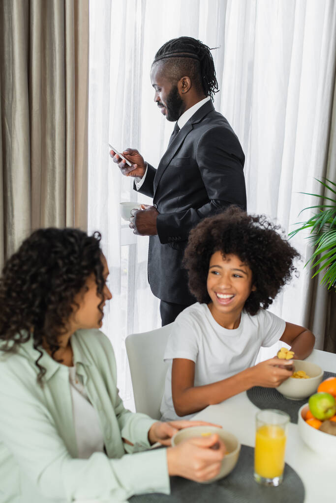 Afro-Amerikan takım elbiseli adam akıllı telefon kullanıyor ve ailesinin yanında kahve tutuyor bulanık ön planda kahvaltı yapıyor.  - Fotoğraf, Görsel
