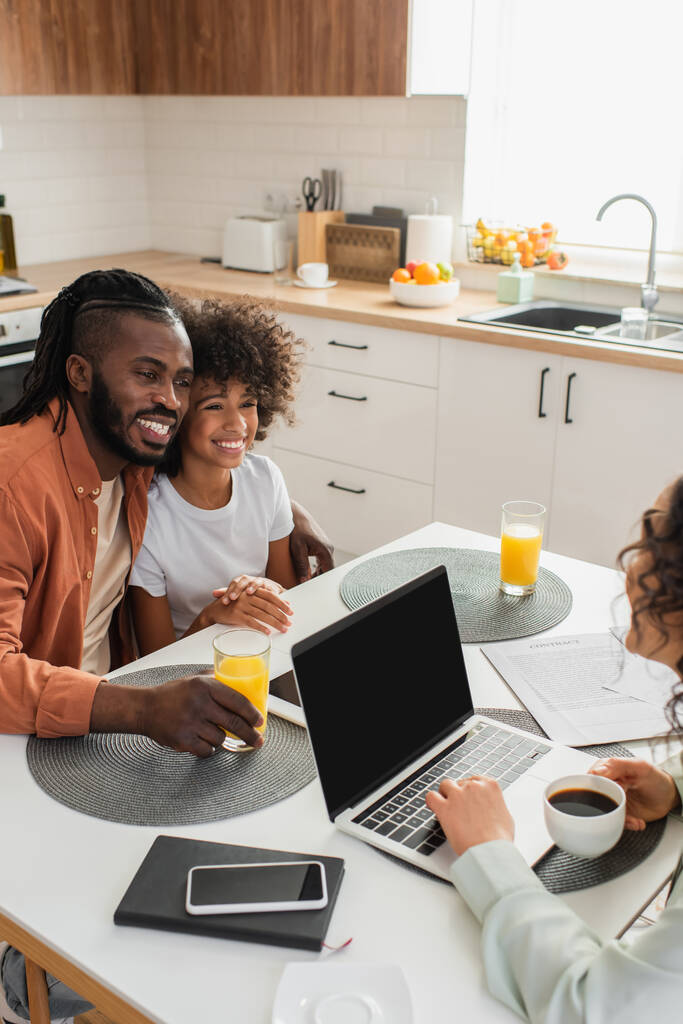 donna afro-americana che tiene la tazza vicino al computer portatile mentre marito e figlia felici sorridono in cucina  - Foto, immagini