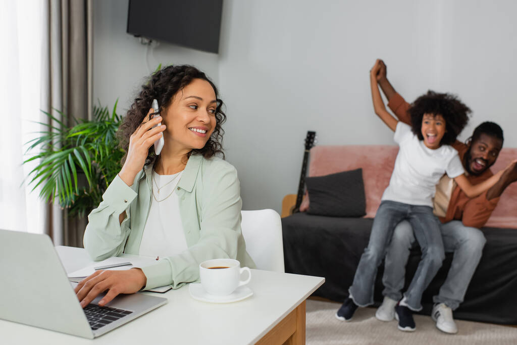 veselý africký Američan žena mluví na smartphone a pomocí notebooku v blízkosti šťastné rodiny na rozmazaném pozadí - Fotografie, Obrázek