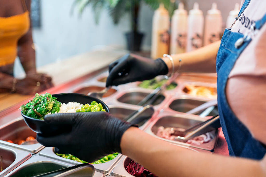 Μη αναγνωρισμένος υπάλληλος εστιατορίου με προστατευτικά γάντια που προσθέτει λαχανικά σε σακουλάκι. - Φωτογραφία, εικόνα