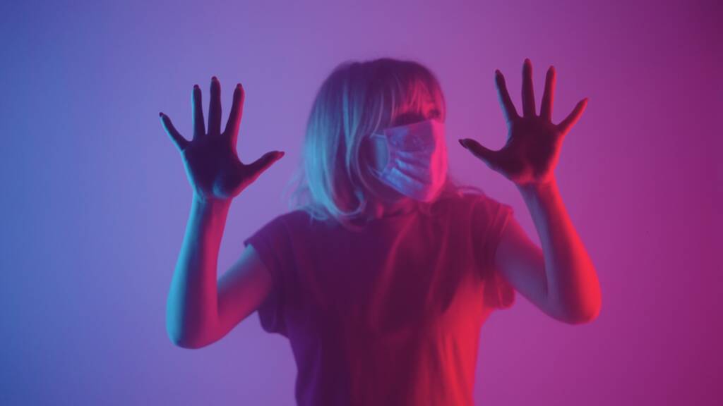 医療用マスクの女性側に見て、ガラスの上で彼女の手のひらに触れる。紫のピンクの柔らかい光と煙を背景に暗いスタジオで手で動揺した女性がガラスに触れる。隔離 - 写真・画像