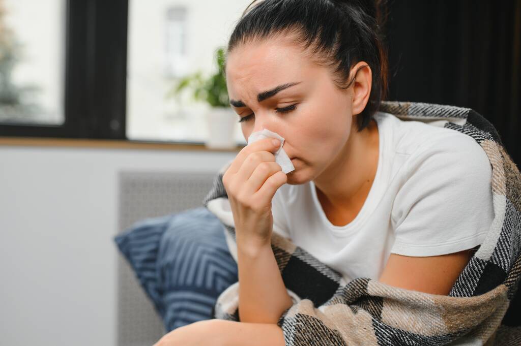 Mala joven sentada en un sofá cubierto de manta, congelada, sonándose la nariz mocosa, estornudando en tejido. La mujer tiene fiebre, gripe y síntomas de gripe tos en casa. Enfermo alérgico con enfermedad respiratoria. - Foto, imagen