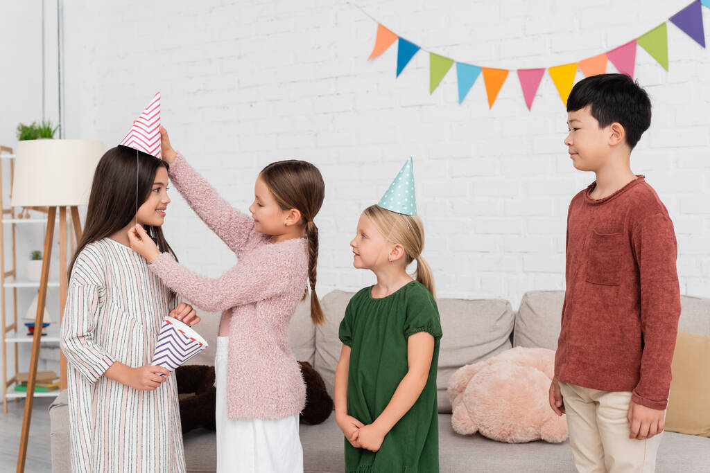 Smiling girl wearing party cap on friend near interracial kids at home  - Photo, Image